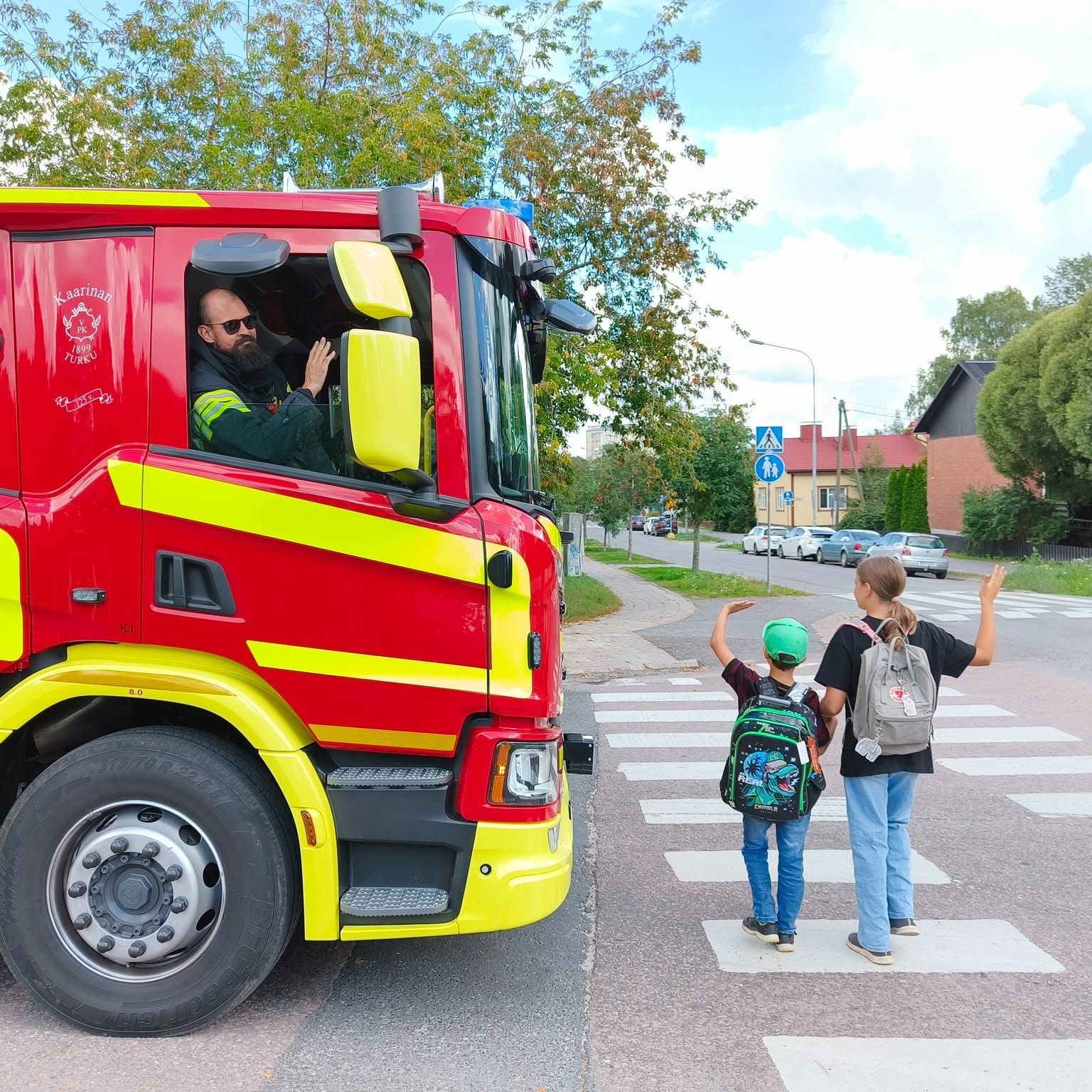 Kaarinan VPK:n paloauton kuljettaja vilkuttaa kahdelle repullaan koululaiselle suojatiellä.